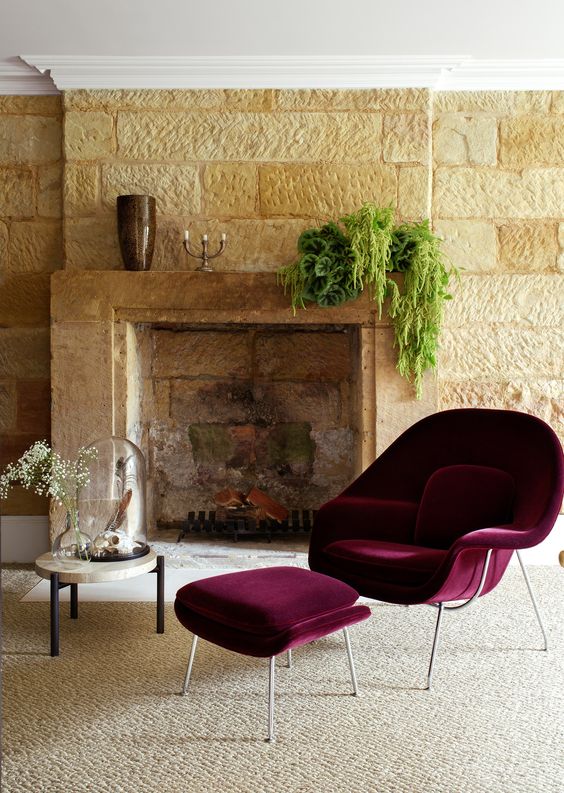 a sophisticated living room with vintage stone cladding, a mantel with greenery, a burgundy Wumb chair with an ottoman and a side table