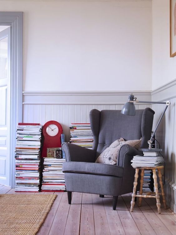 a vintage reading nook with a grey Strandmon chair, a wooden stool, stacked books and magazines and a grey floor lamp
