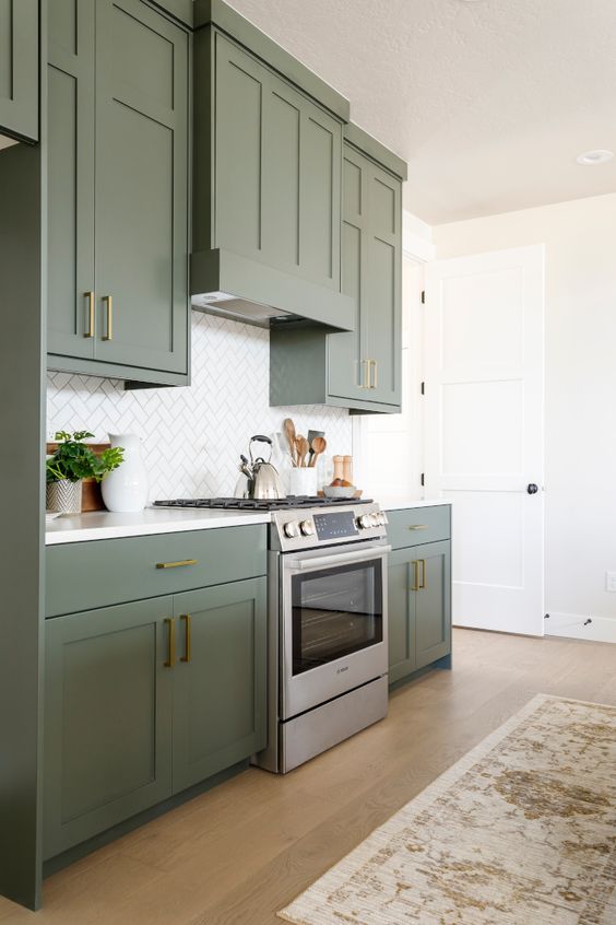 an elegant olive green kitchen with shaker cabinets, gold and brass handles, a white tile backsplash and a printed rug is cool