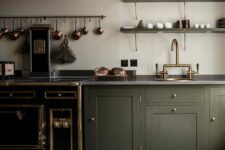 an elegant vintage olive green kitchen with shaker cabinets, grey stone countertops, a large hood, copper and brass touches