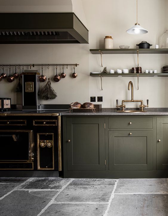 an elegant vintage olive green kitchen with shaker cabinets, grey stone countertops, a large hood, copper and brass touches