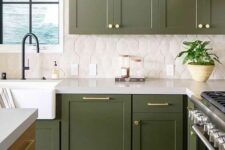 an olive green farmhouse kitchen with shaker cabinets, a catchy white tile backsplash, black fixtures and gold handles and knobs
