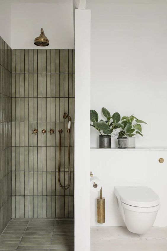 a chic modern bathroom with a shower space clad with olive green skinny tiles, a white space with a toilet and potted plants