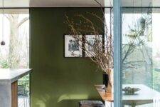 03 a cool dining space with an olive green accent wall, a wooden trestle dining table, a wooden bench and some branches in a vase