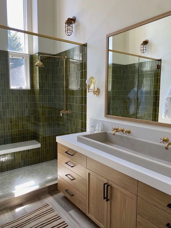 a farmhouse bathroom with a large shower space with olive green skinny tiles, a large wooden vanity, a large mirror and gold and black fixtures