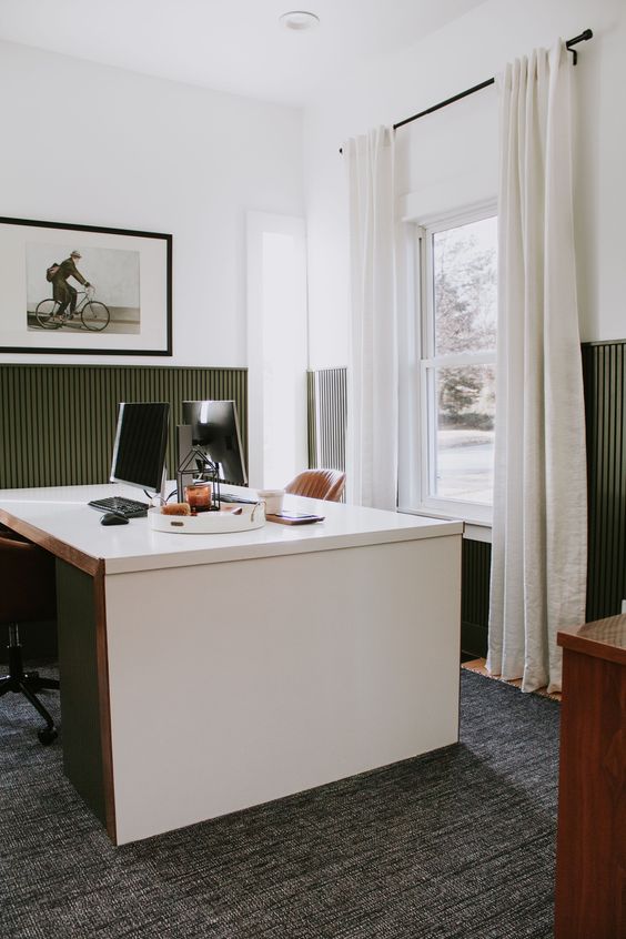a farmhouse home office with olive green paneling, a shared white desk, a large rug, amber leather chairs and some art
