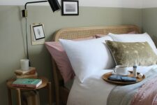 15 a pretty and welcoming bedroom with white and olive green walls, a rattan bed and neutral and green bedding, a wooden nightstand and a black sconce