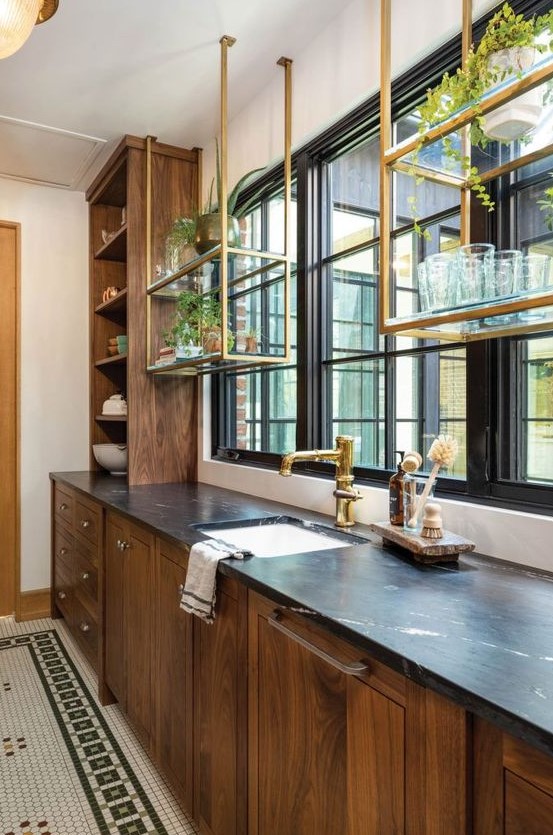 a stained kitchen with black stone countertops and brass suspended shelves over the windows is a cool and elegant space