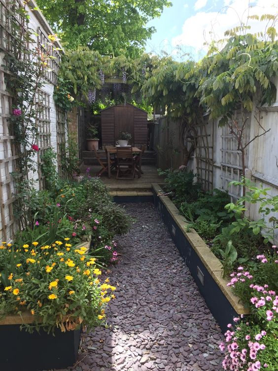 a long and narrow backyard with a gravel path, raised garden beds with greenery and blooms, a deck with a dining space a long and narrow backyard with a gravel path, raised garden beds with greenery and blooms, a deck with a dining space