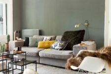 19 a Scandinavian living room with olive green walls, a grey sofa and colorful pillows, a gold sconce, a rocker and some coffee tables