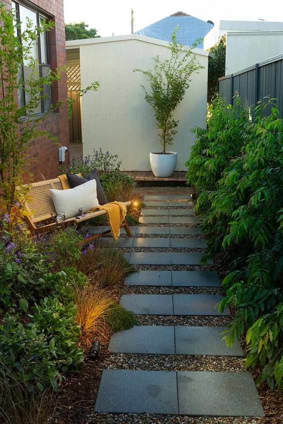 a long and narrow garden with a tile pathway, greenery, grasses and blooms, a rattan bench with pillows and some lights a long and narrow garden with a tile pathway, greenery, grasses and blooms, a rattan bench with pillows and some lights