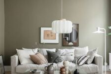 22 an airy living room with olive green walls, a neutral sofa and pillows, a glass coffee table, a pendant lamp and some art