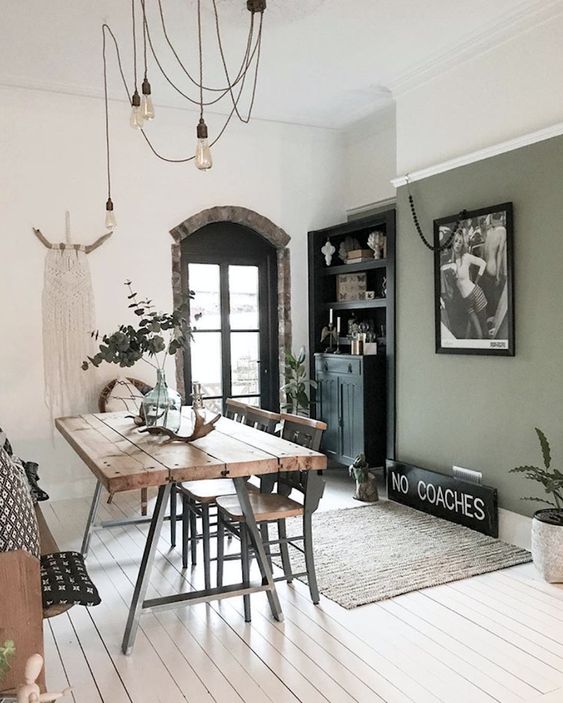 an eclectic dining room with an olive green accent wall, a stained table and metal and wood chairs, hanging bulbs, a built in sideboard