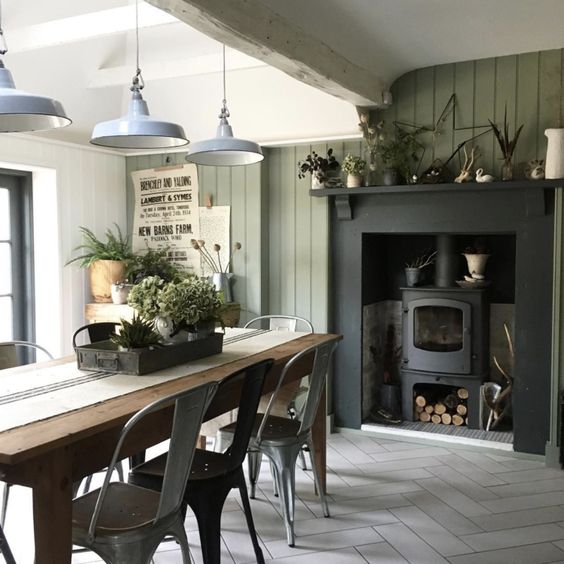 an industrial farmhouse dining room with an olive green shiplap wall, a hearth, a wooden table and metal chairs, metal pendant lamps