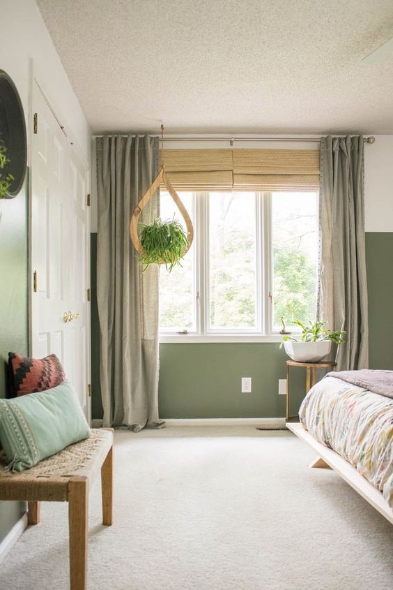 an olive green boho bedroom with a bed with printed bedding, a woven bench and pillows, a potted plant and lovely curtains