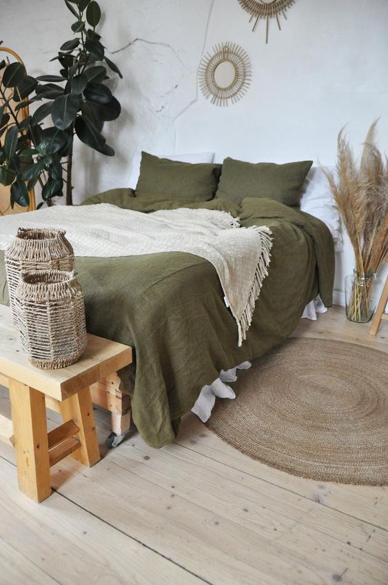 a boho bedroom in neutrals, with a bed done with olive green bedding, a wooden bench and woven candleholders, pampas grass, potted plants and burst mirrors