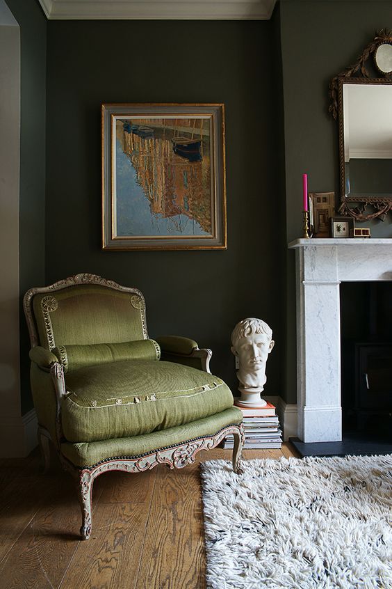 a dark green living room with a fireplace, an olive green chair, some art, books and a mirror in an ornated frame