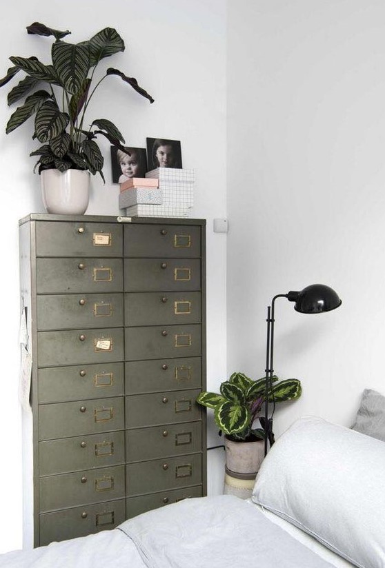 a Scandinavian bedroom with a bed with neutral bedding, a table lamp and some plants, a green card cabinet as a storage unit