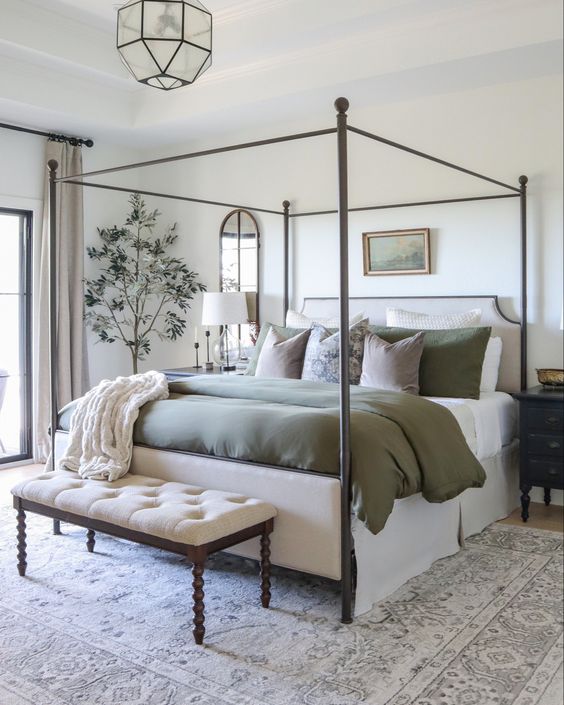 a farmhouse bedroom in neutrals, with a metal frame bed and olive green bedding, an upholstered bench, some art and plants