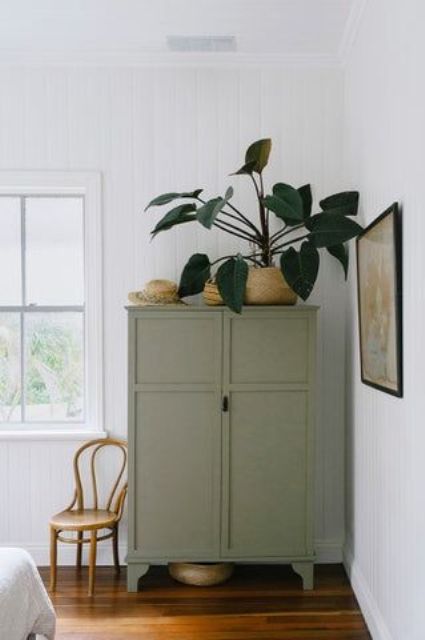 a large and pretty olive green wardrobe, potted plants on top, a chair and a frame artwork for a lovely space