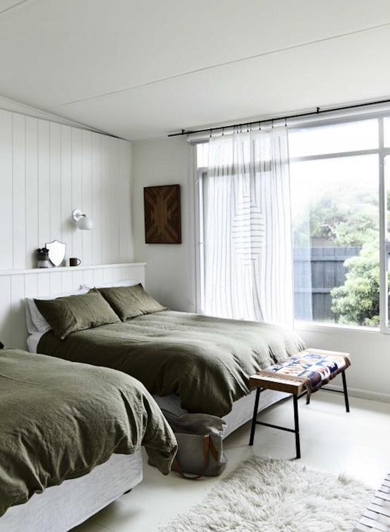 a neutral farmhouse bedroom with white shiplap walls, two beds, olive green bedding, an upholstered bench and a fluffy rug