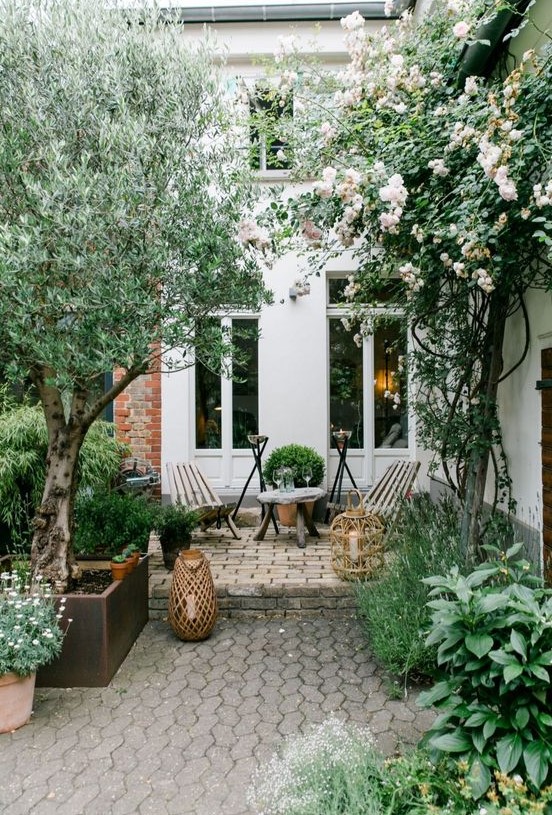 a simple modern paved backyard with wooden furniture, some candles in large candle lanterns and trees and blooms a simple modern paved backyard with wooden furniture, some candles in large candle lanterns and trees and blooms