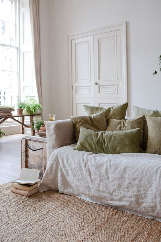 a neutral living room with a neutral sofa, olive green pillows, a wooden chest for storage and a bench by the window
