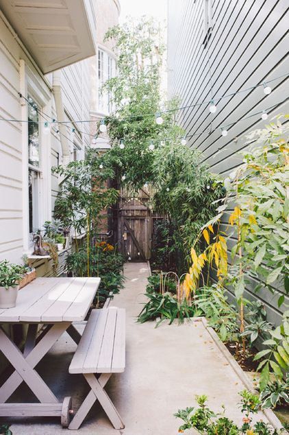 a small side garden space clad with concrete, wiht greenery and plants, with a whitewashed dining set and lights over the space a small side garden space clad with concrete, wiht greenery and plants, with a whitewashed dining set and lights over the space