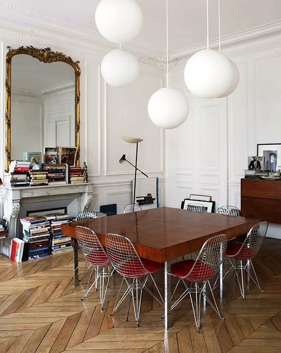 a Parisian dining room with a fireplace filled with books, a large mirror in a gilded frame, a stained storage unit, a stained table and Eames wire chairs plus pendant bubble lamps
