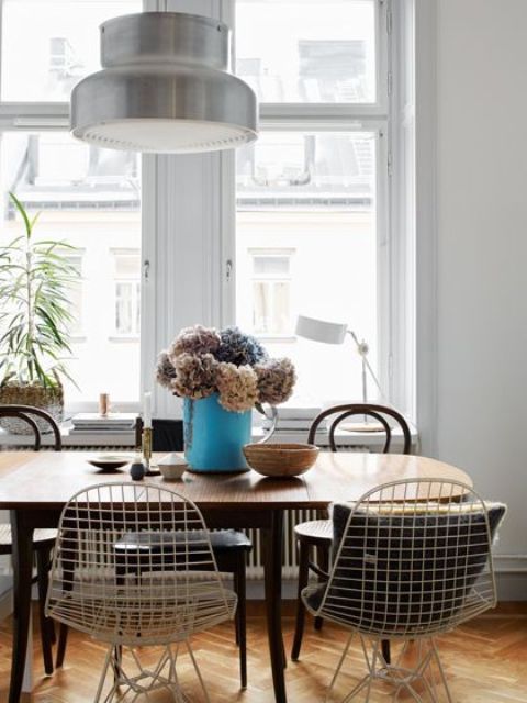 a Scandinavian dining room with an oval stained table, metal Eames wire chairs, a metal pendant lamp and some lovely blooms and plants