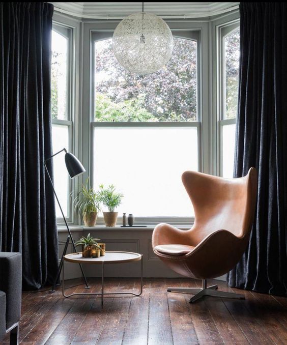 a bay window with some plants and black curtains, an amber leather Egg chair, a side table and a black floor lamp