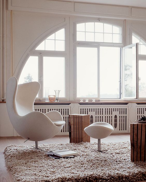 a beautiful neutral nook with an arched window, a creamy Egg chair and a footrest, wooden stools, a fluffy rug and some decor