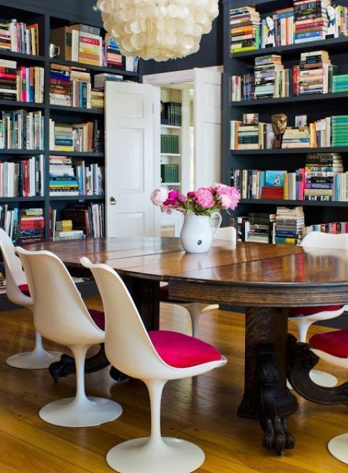a bold dining room with graphite grey walls and storage units with books, a large stained table, pink Tulip chairs and a seashell chandelier