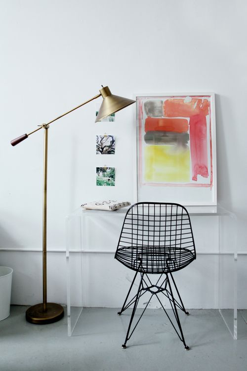 a bold space with a clear acrylic desk, a black Eames wire chair, bold artwork and a brass floor lamp is all cool