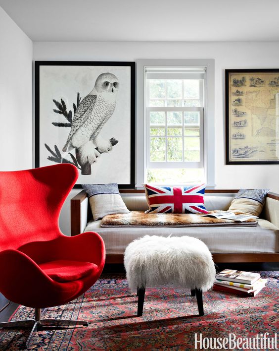 a bright living room with oversized artwork, a grey sofa with bold pillows and blankets, a bold red Egg chair, an ottoman and a printed rug