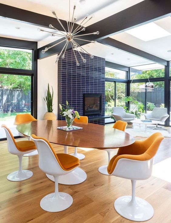 a bright mid-century modern dining room with an oval table and rust-colored tulips chairs, a fireplace clad with navy skinny tiles and a sunburst chandelier