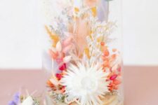 a bright pastel dried flower arrangement in pink, orange, lilac, red and neutral blooms and grasses in a neutral cloche