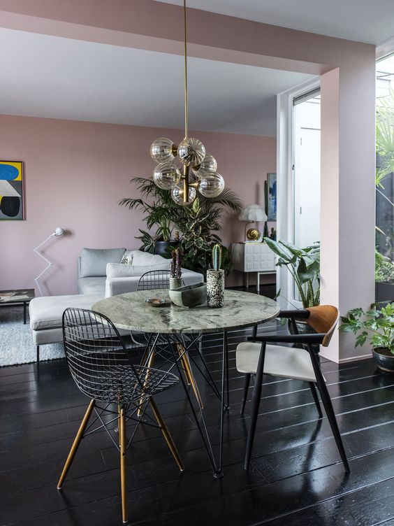 a bright space with pink walls, a grey sofa, a round table, mismatching chairs including Eames wire ones and a catchy chandelier