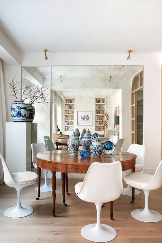 a chic dining room with a mirror accent wall, a stained table and white Tulip chairs, a storage unit with a blue vase and some vases on the table