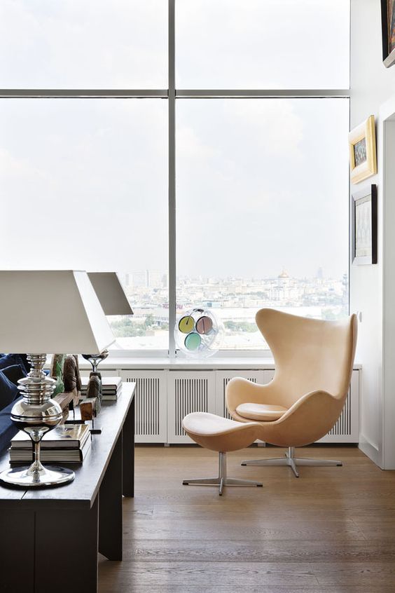 a chic nook by the window, with a warm-colored leather Egg chair and a footrest, a gallery wall and a console table with books and lamps