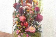 a cloche with a wooden base, with moss, bold pink and red dried blooms, leaves and some grasses is a colorful summer home decoration