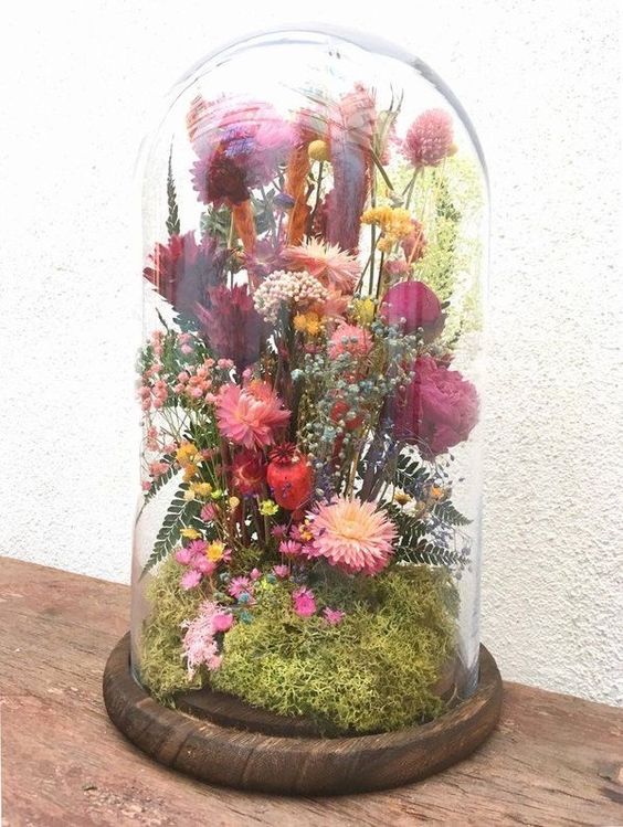 a cloche with a wooden base, with moss, bold pink and red dried blooms, leaves and some grasses is a colorful summer home decoration