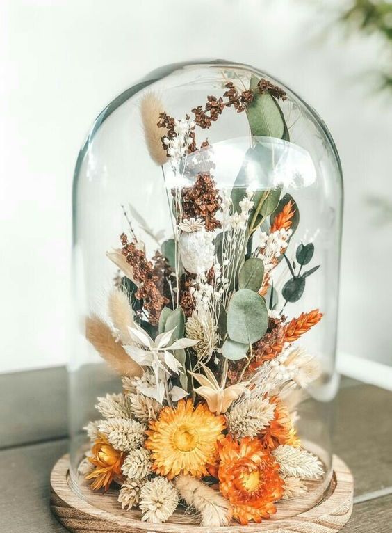 a cloche with a wooden base, with orange dried blooms, grasses, greenery feels like summer and can be a nice decoration for the fall, too