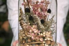 a cloche with gilded acorns, dried blooms, dark grasses and gilded branches and some pink flowers is a lovely decoration for the fall
