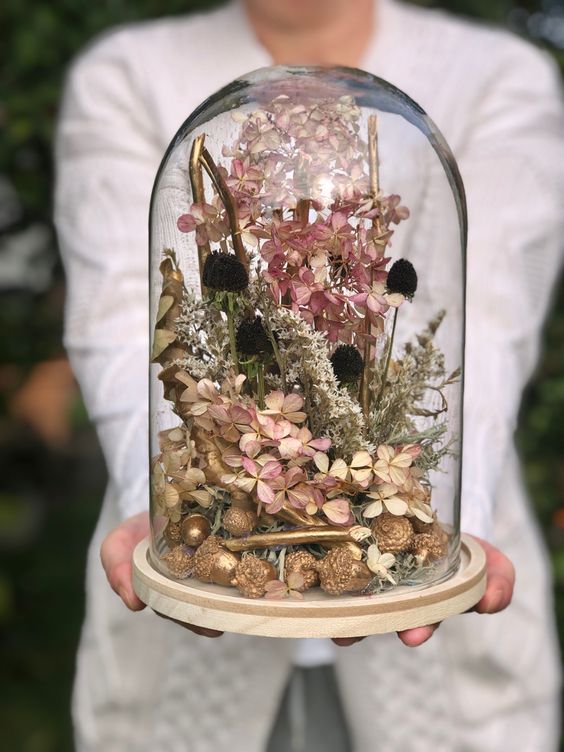 a cloche with gilded acorns, dried blooms, dark grasses and gilded branches and some pink flowers is a lovely decoration for the fall
