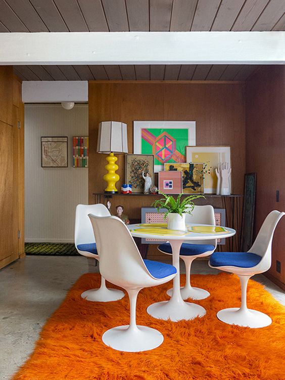 a colorful dining room with stained walls, a console table with a gallery wall, a round table, bold blue Tulip chairs, an orange rug