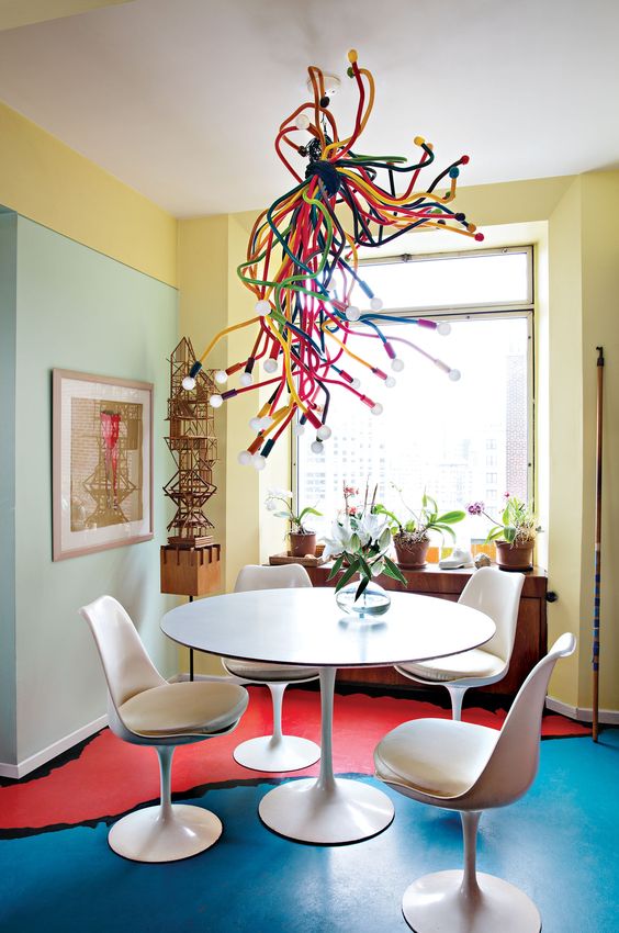 a colorful dining space with color block walls and a floor, a round table and neutral Tulip chairs and a colorful bulb chandelier