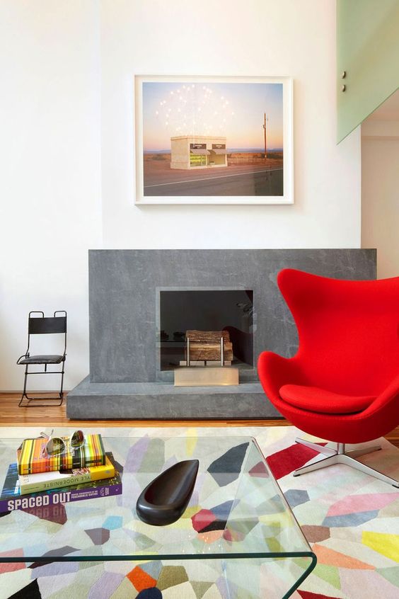 a colorful living room with a fireplace, a red Egg chair, a colorful printed rug, a glass coffee table, a bold photo