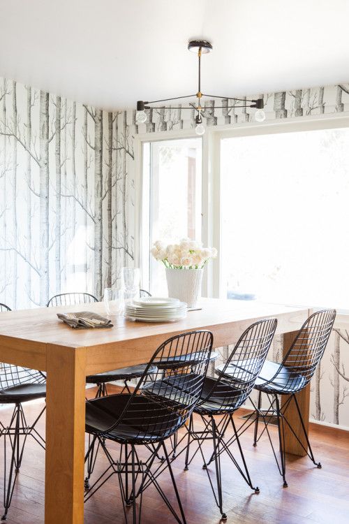a cozy dining room with wood print wallpaper, a stained table, black Eames wire chairs and a chandelier plus some blooms