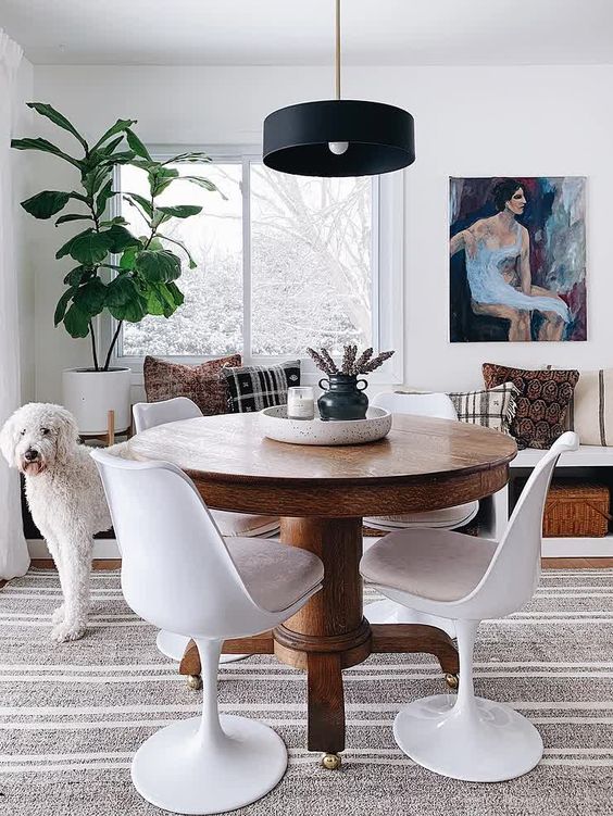 a cozy dining space with a windowsill bench with printed pillows, a stained table, blush Tulip chairs, a black pendant lamp